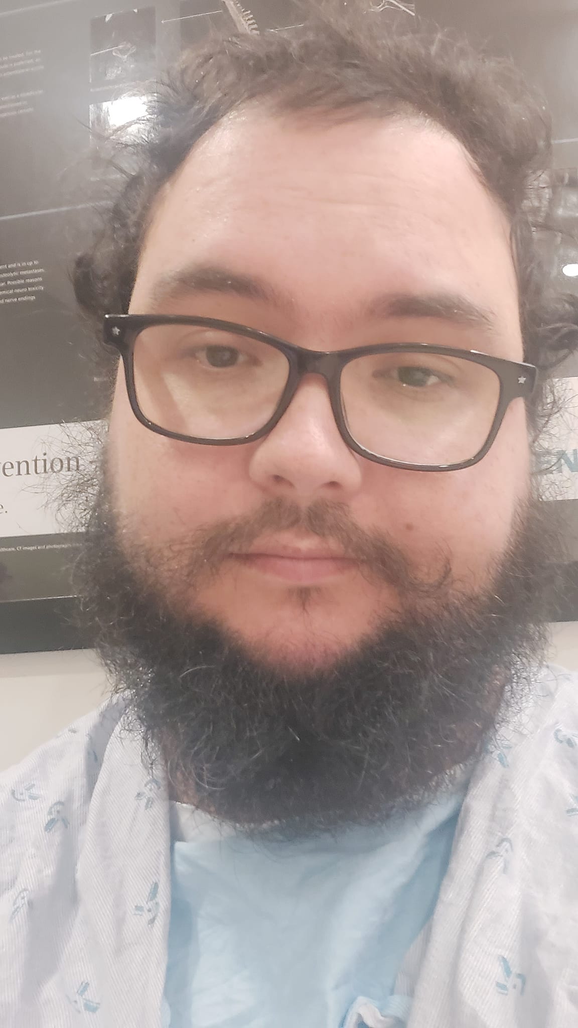 A man with short hair and bushy facial hair sits in a different hospital waiting room, wearing a hospital gown